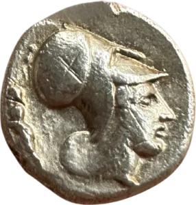 Tête d'Athéna à droite coiffée d'un casque Corinthien. Un symbole (guerrier?) derrière la tête.